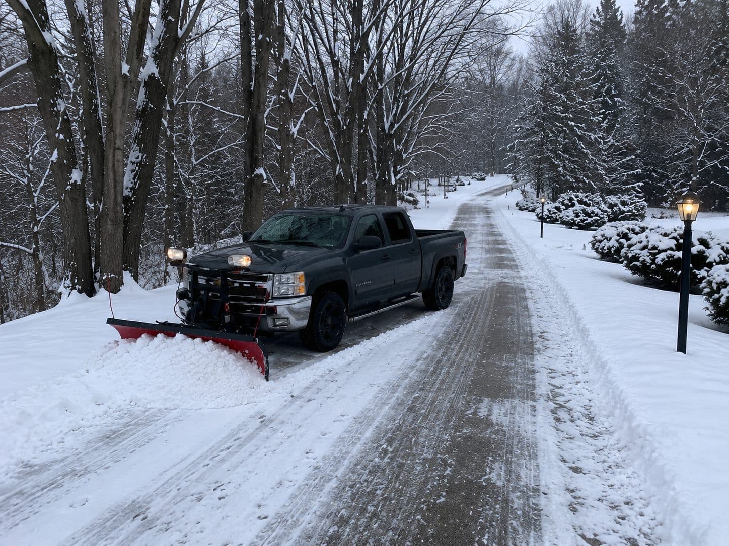 A Fresh Cut Lawns truck with a red plow blade clearing snow from a tree-lined residential driveway in winter